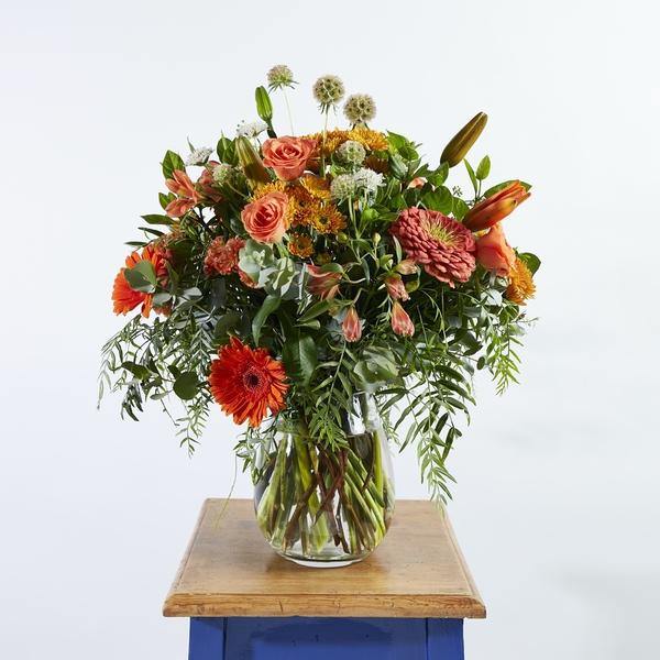 Orange roses, lilies, gerberas arranged with whimsical greenery in a glass vase