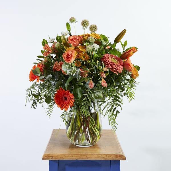 Orange roses, lilies, gerberas arranged with whimsical greenery in a glass vase